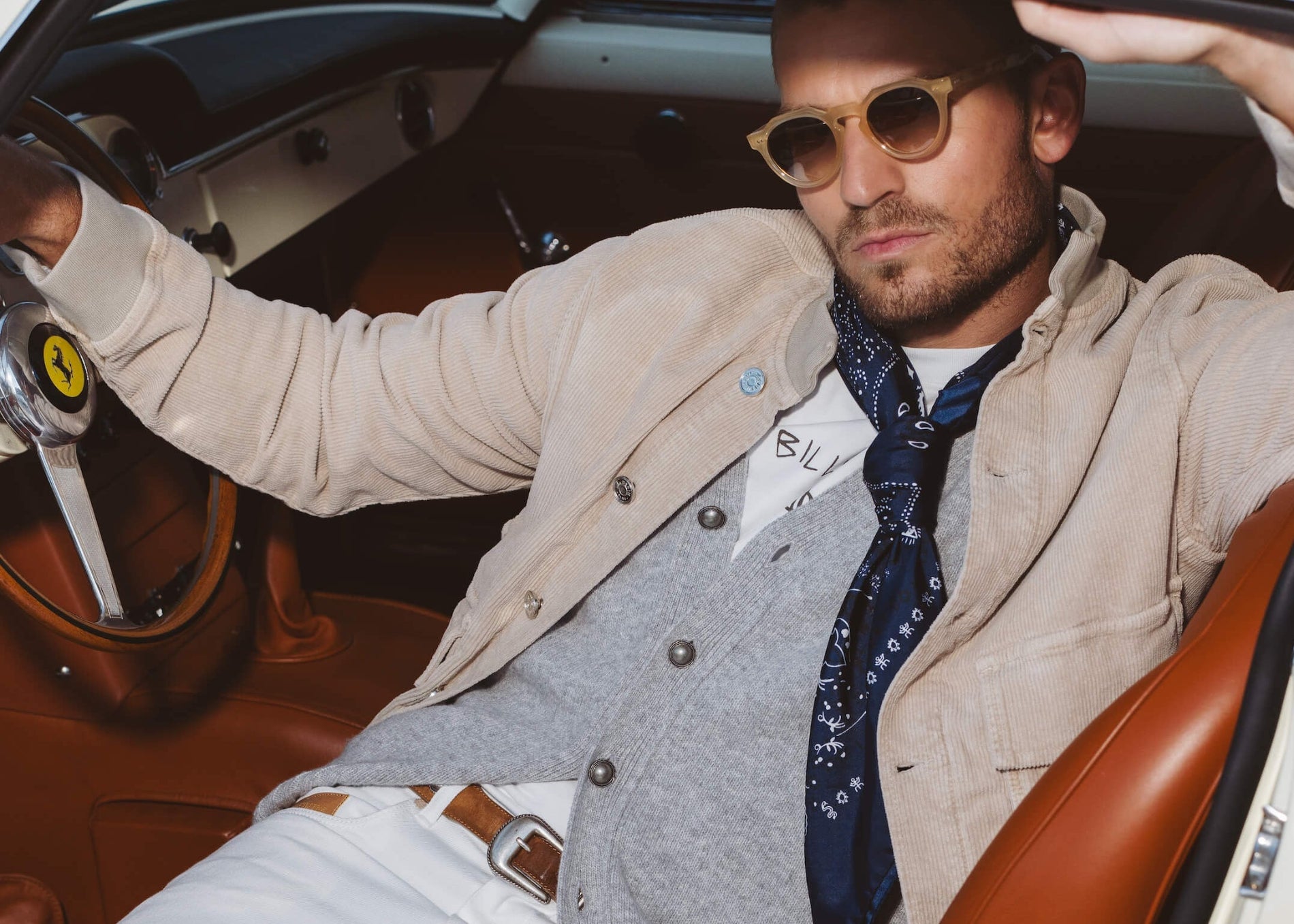 Mannequin wearing a grey cardigan, white jeans, a velvet jacket, and a brown belt sitting in a classic Ferrari